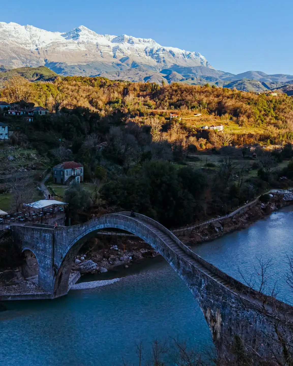 Aerial photograph of a stone bridge, river and mountain village by Dimitris Kyriakoulis