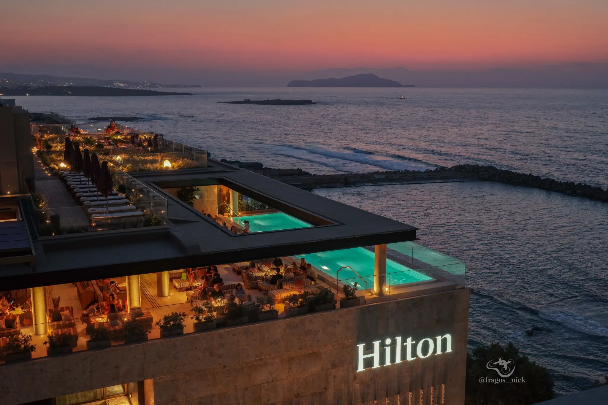 Aerial dusk photograph of a seafront Hilton hotel and illuminated pools by Nikos Fragos