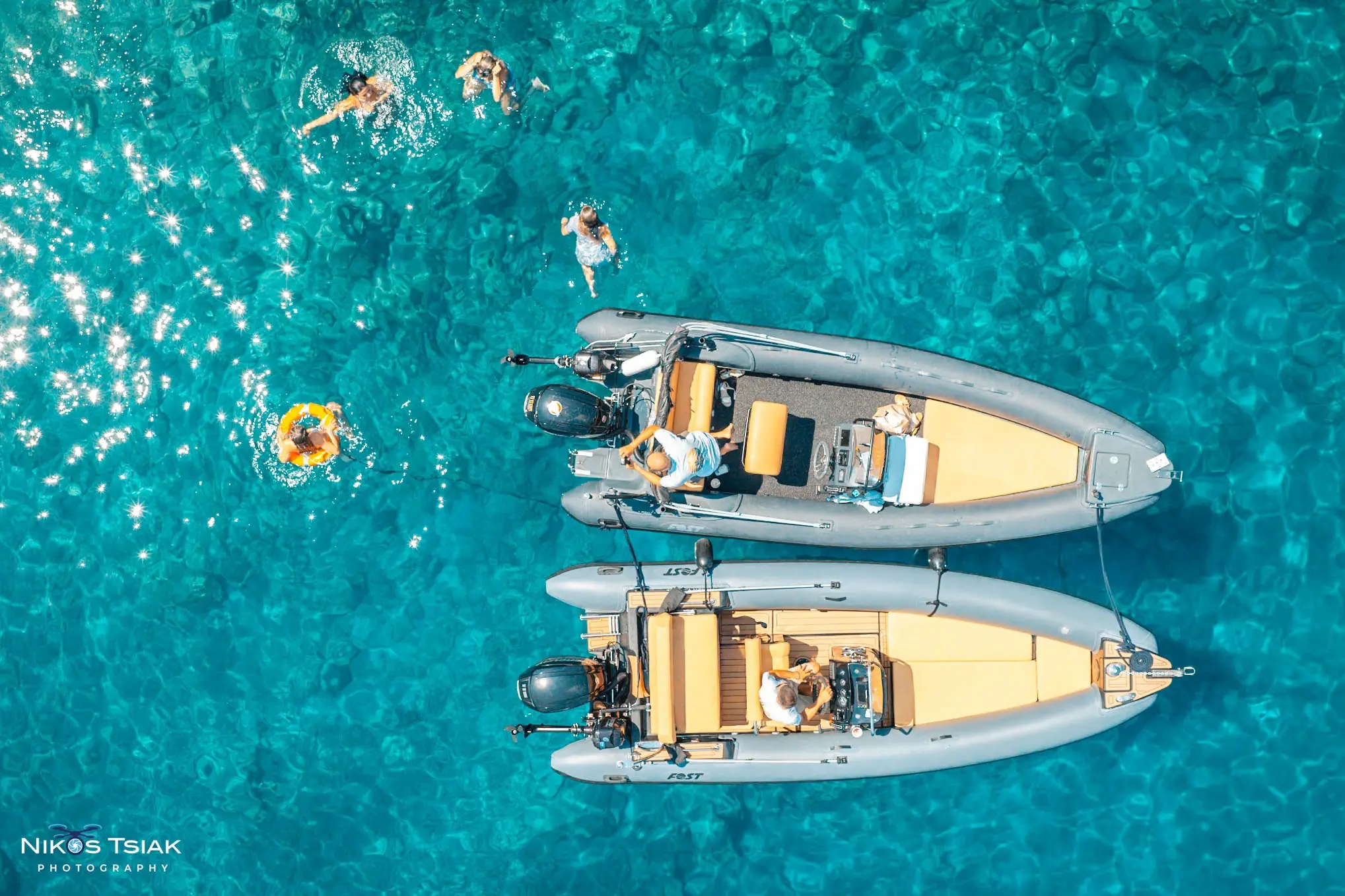 Top-down aerial photograph of two boats and swimmers in clear turquoise water by Nikos Tsiakalakis