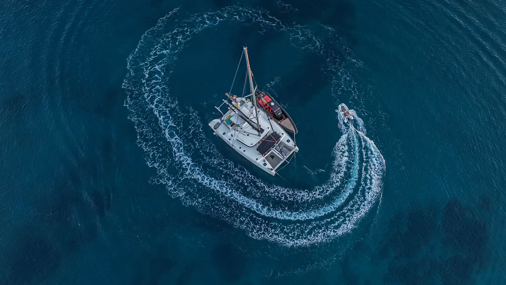 Top-down aerial photograph of a catamaran and a speedboat circling in deep blue water by Themistoklis Karantinos